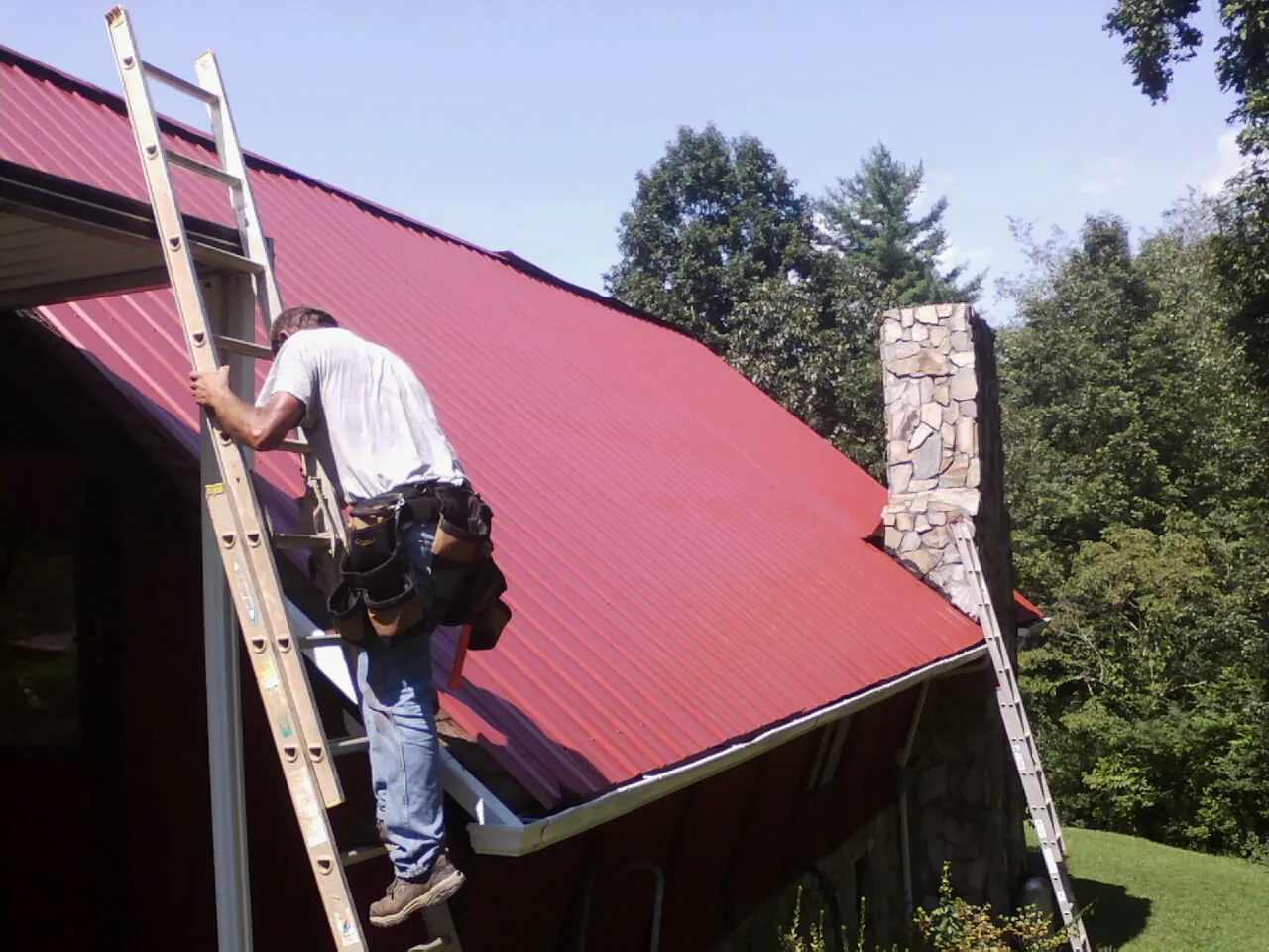 Roofer on a ladder beside a red metal roof and stone chimney — Soffit & Fascia Repair by Roofing Brothers in Basalt